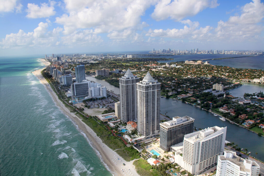 Shows the islands of Miami.  In the front is tall condo buildings of the beach, in the rear skyscrapers of downtown Miami Skyline.  Blue Atlantic ocean waves going to the beach.